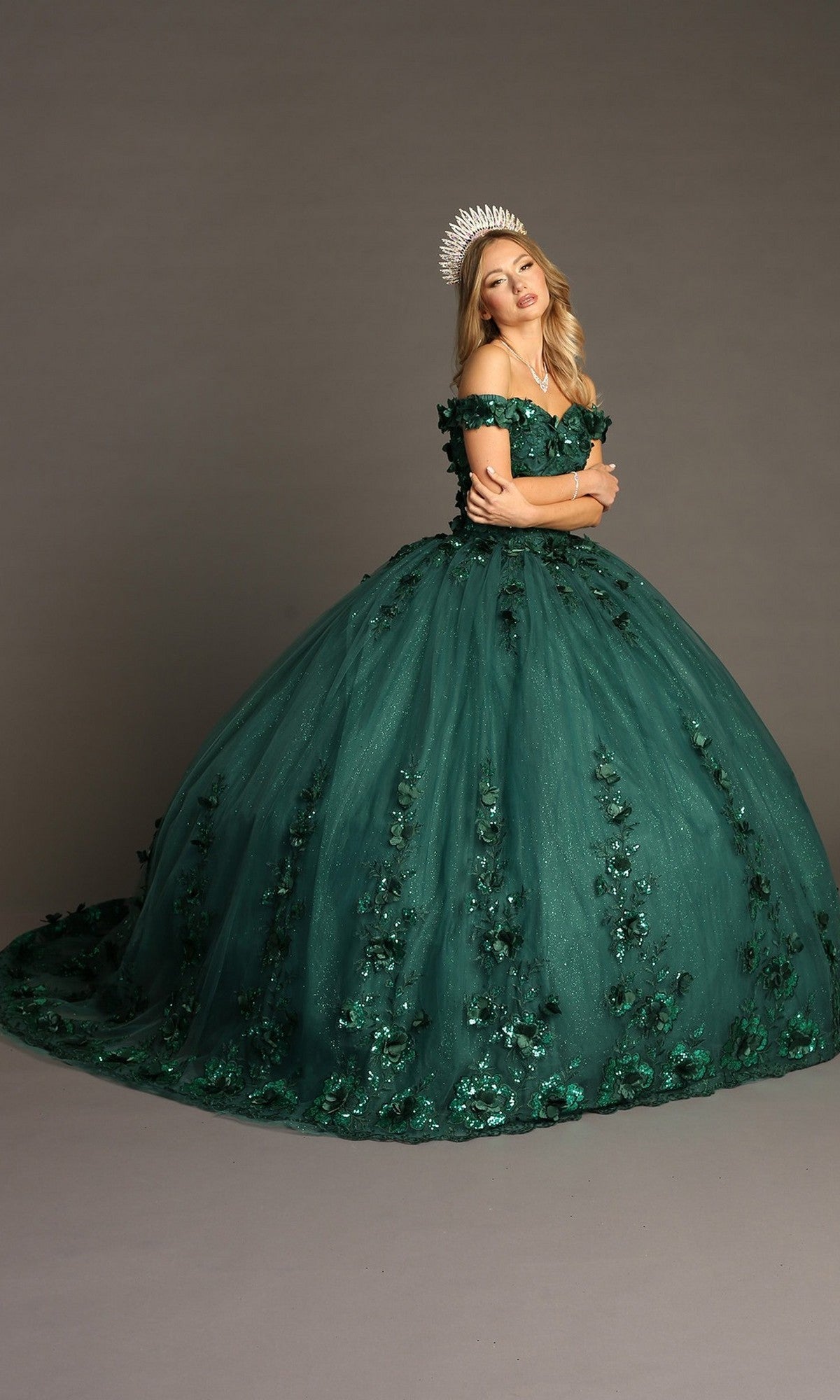 A woman in the Quinceanera Dress CHF3082 By Chicas, a dark green glitter tulle ballgown adorned with 3D floral appliques and elegant off-the-shoulder styling, stands against a plain background. She completes the look with a white crown, highlighting the gownโs intricate floral details throughout.